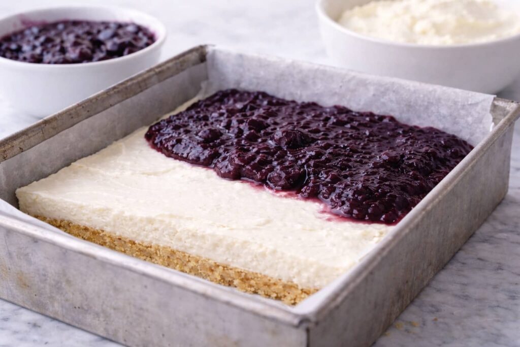 Blueberry cream cheese bars layering process in square pan showing crust, cream cheese filling, and partially spread blueberry topping