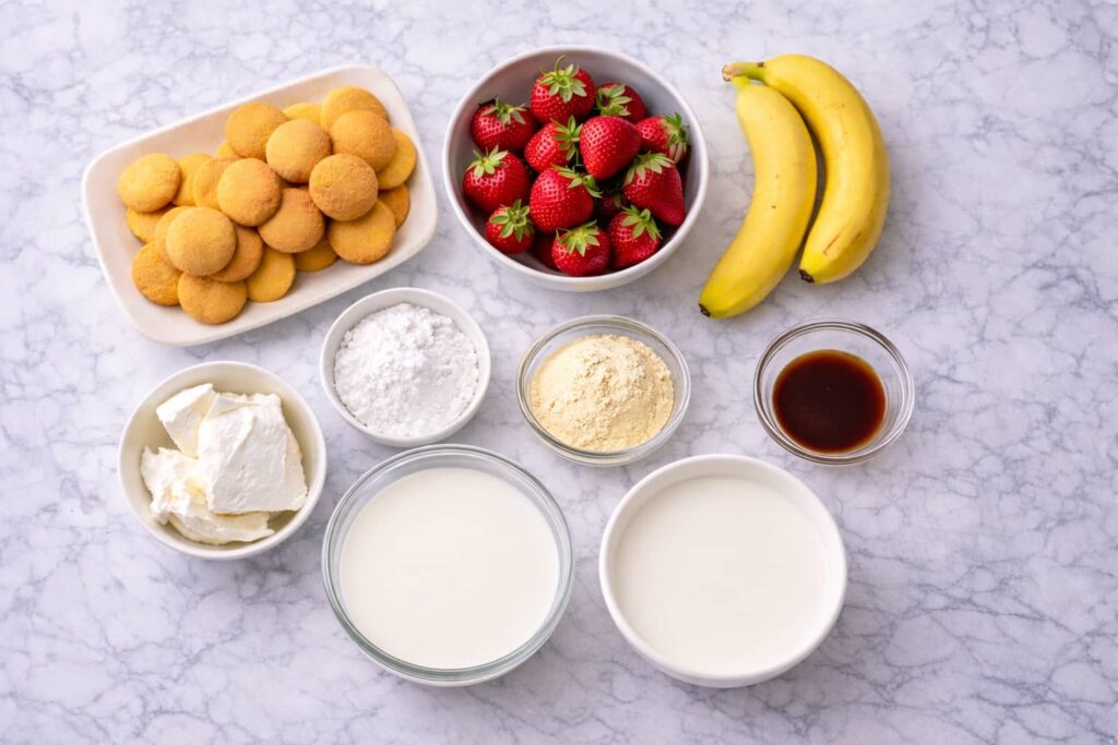 Ingredients for strawberry cheesecake banana pudding arranged on white marble surface