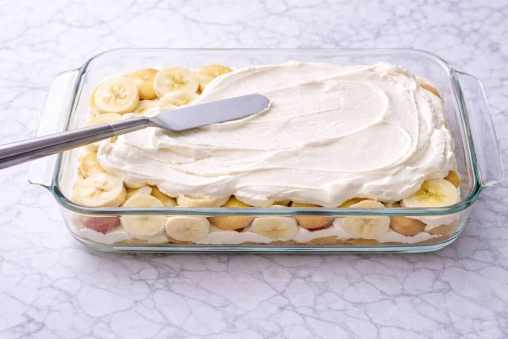 Spreading cheesecake layer over bananas and vanilla wafers in glass baking dish