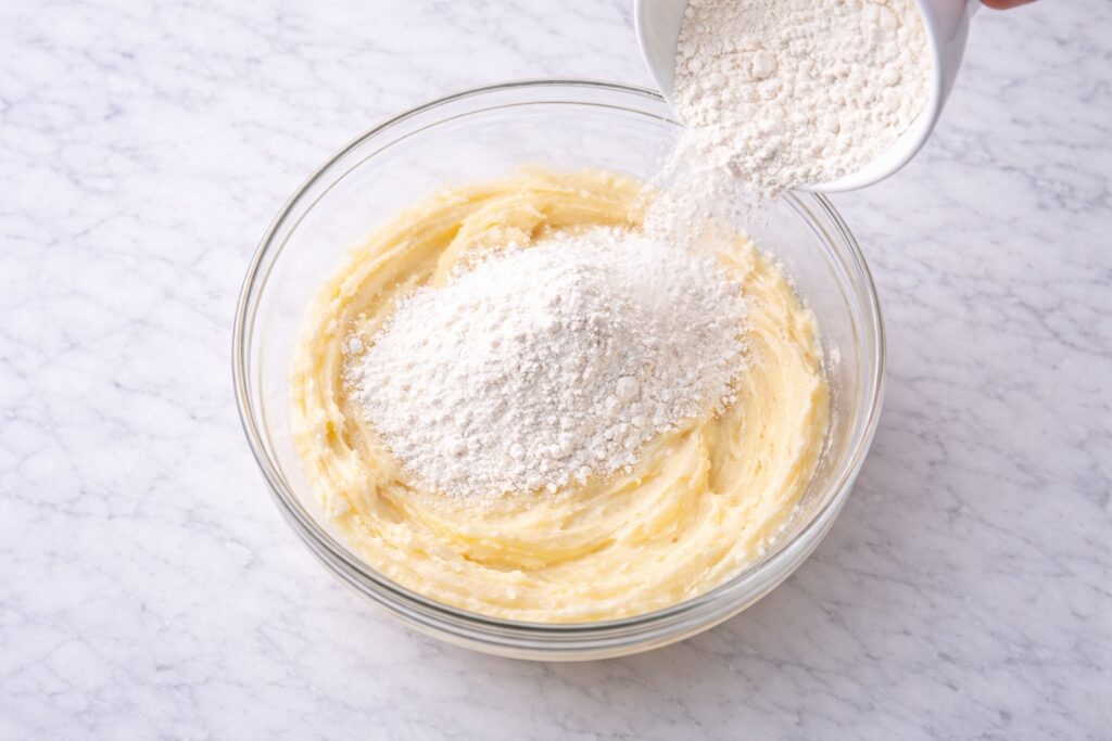 Flour being added to lemon cake batter in a glass bowl to begin mixing the dry ingredients