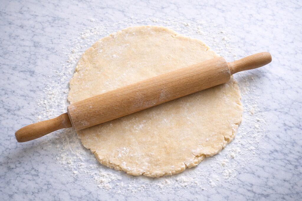 Rolling pie dough on a floured marble countertop with wooden rolling pin