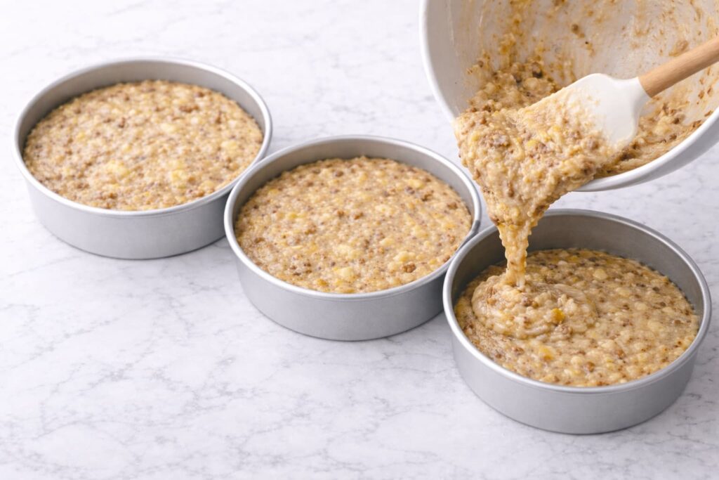 Dividing Hummingbird Cake Batter Evenly Between Pans Hummingbird cake batter being evenly poured into three cake pans with visible fruit and nuts