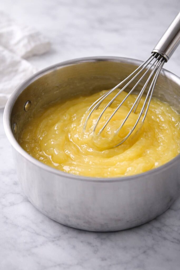 Lemon pie filling thickening in a stainless steel saucepan while being whisked on white marble surface