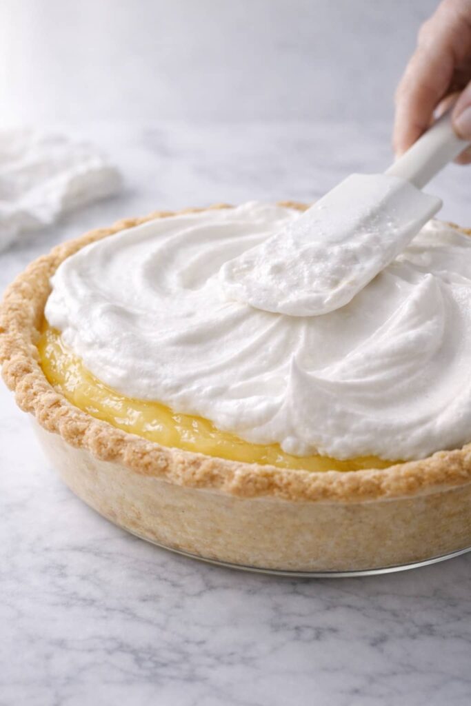 Spreading fluffy meringue over hot lemon filling and sealing edges in pie crust on white marble surface