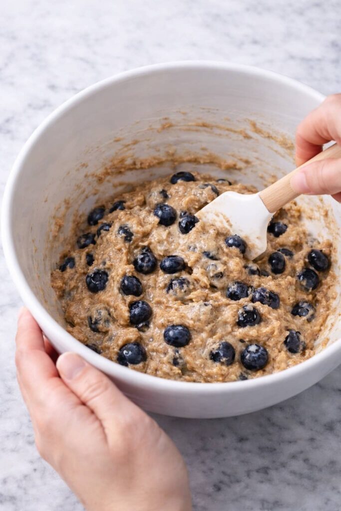 folding fresh blueberries into whole wheat healthy blueberry muffin batter in white mixing bowl