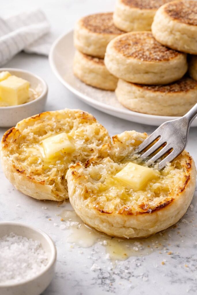 Sourdough English muffins split with a fork, showing airy nooks and crannies with melted butter on a marble surface