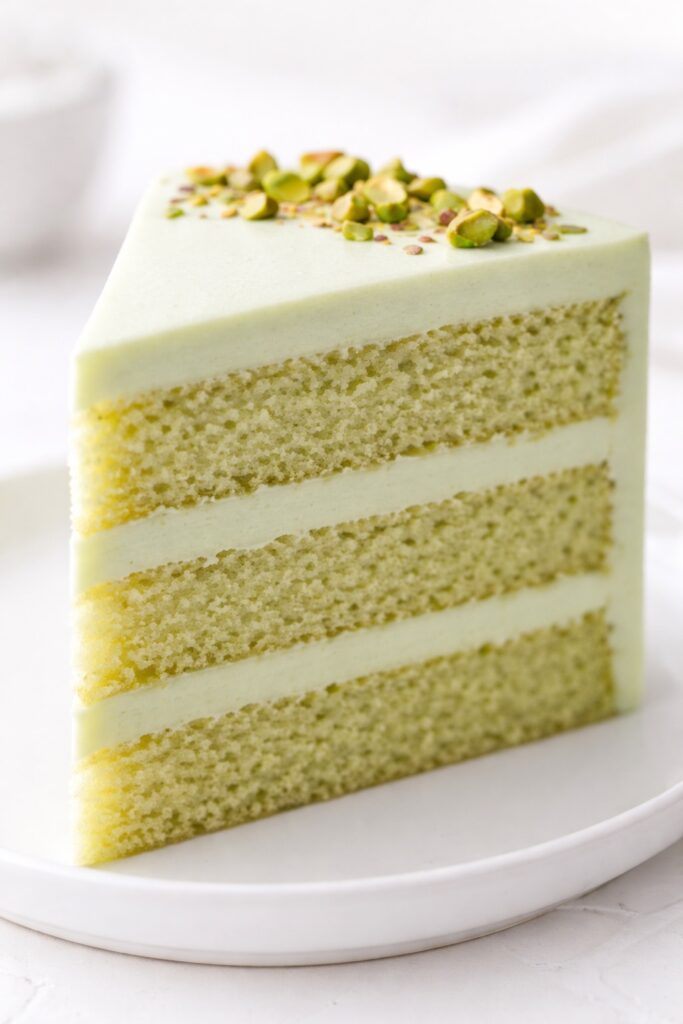 Slice of pistachio layer cake showing three soft cake layers, creamy filling, and smooth pistachio frosting close up