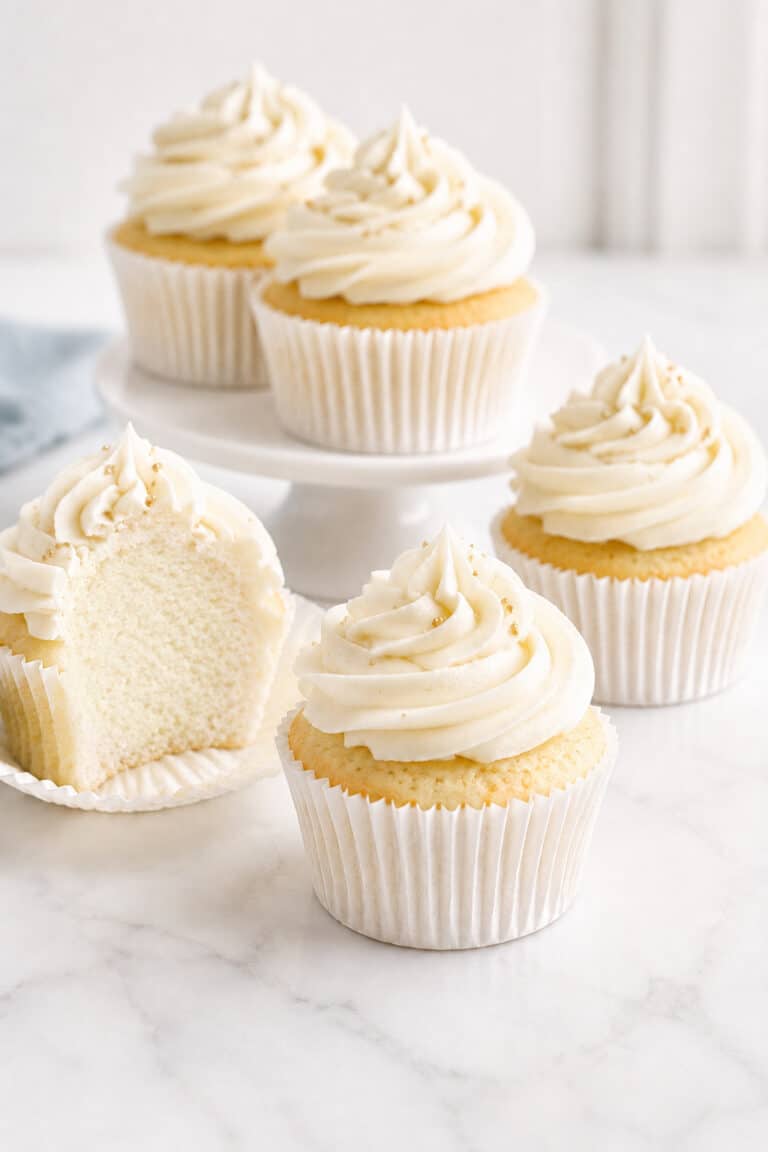 Vanilla cupcakes with pale vanilla buttercream and one cupcake opened to show the soft, fine crumb.