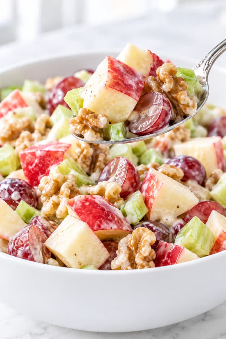 Close-up of apple salad with red-skinned apple cubes, halved red grapes, celery, and walnuts in a white bowl, with one bite lifted on a spoon.