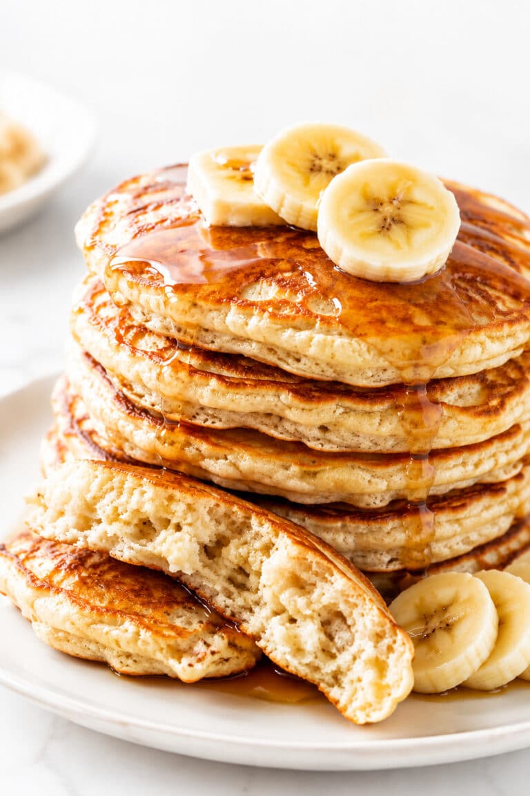 Stack of golden banana pancakes with maple syrup, banana slices, and a broken pancake showing the fully cooked crumb