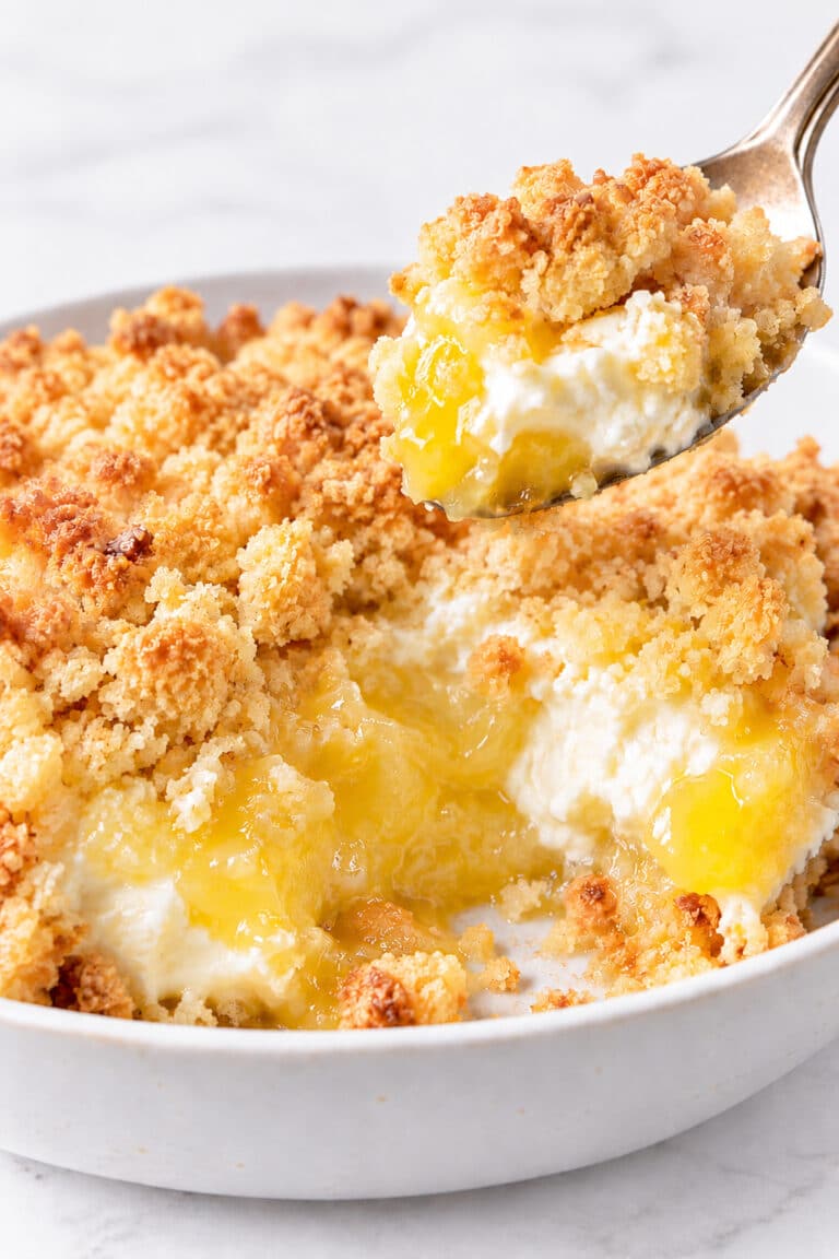 A spoon lifts golden crumb, glossy lemon filling and cream cheese from a white baking dish.