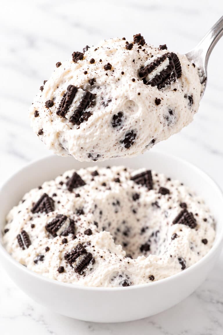 A spoon lifts pale Oreo fluff with dark cookie pieces above a white ceramic bowl.