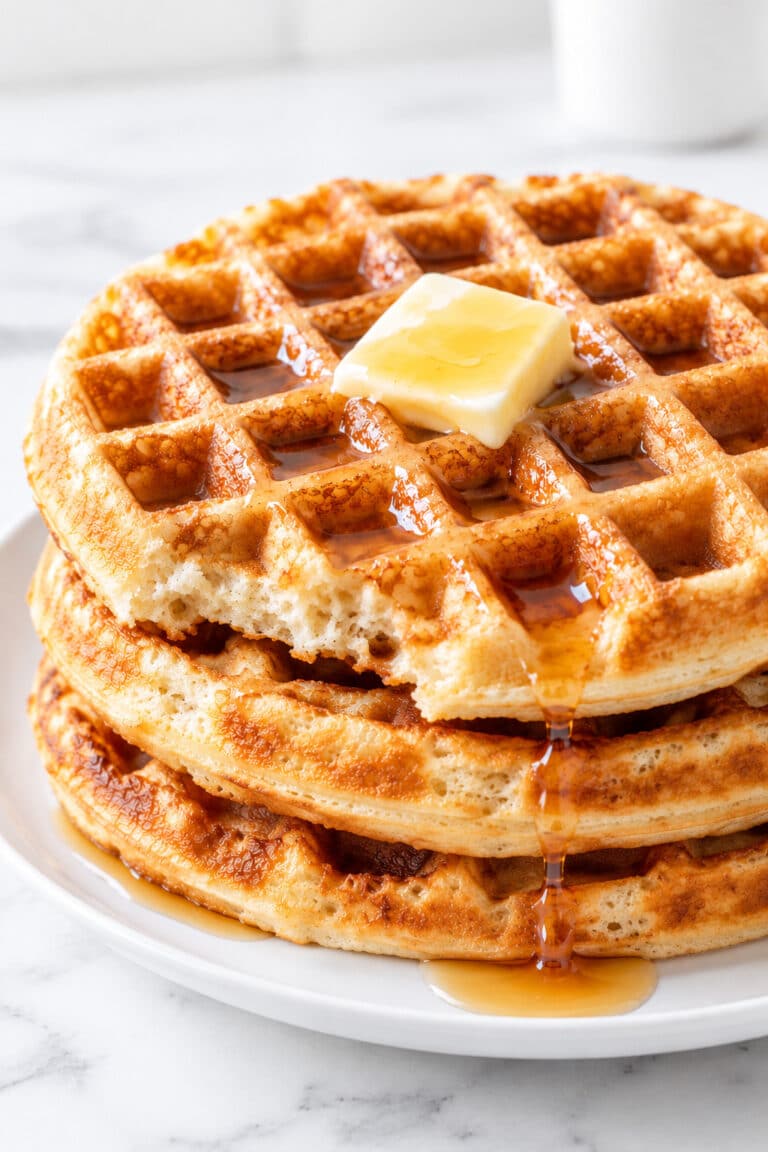 Three golden round sourdough waffles stacked with softened butter and maple syrup on a white plate.