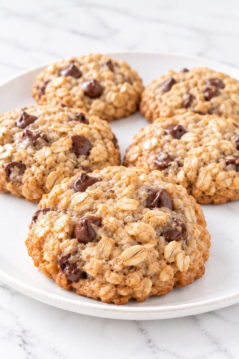 Five whole banana oatmeal cookies on a white plate, with a close foreground cookie showing rolled oats and chocolate chips.